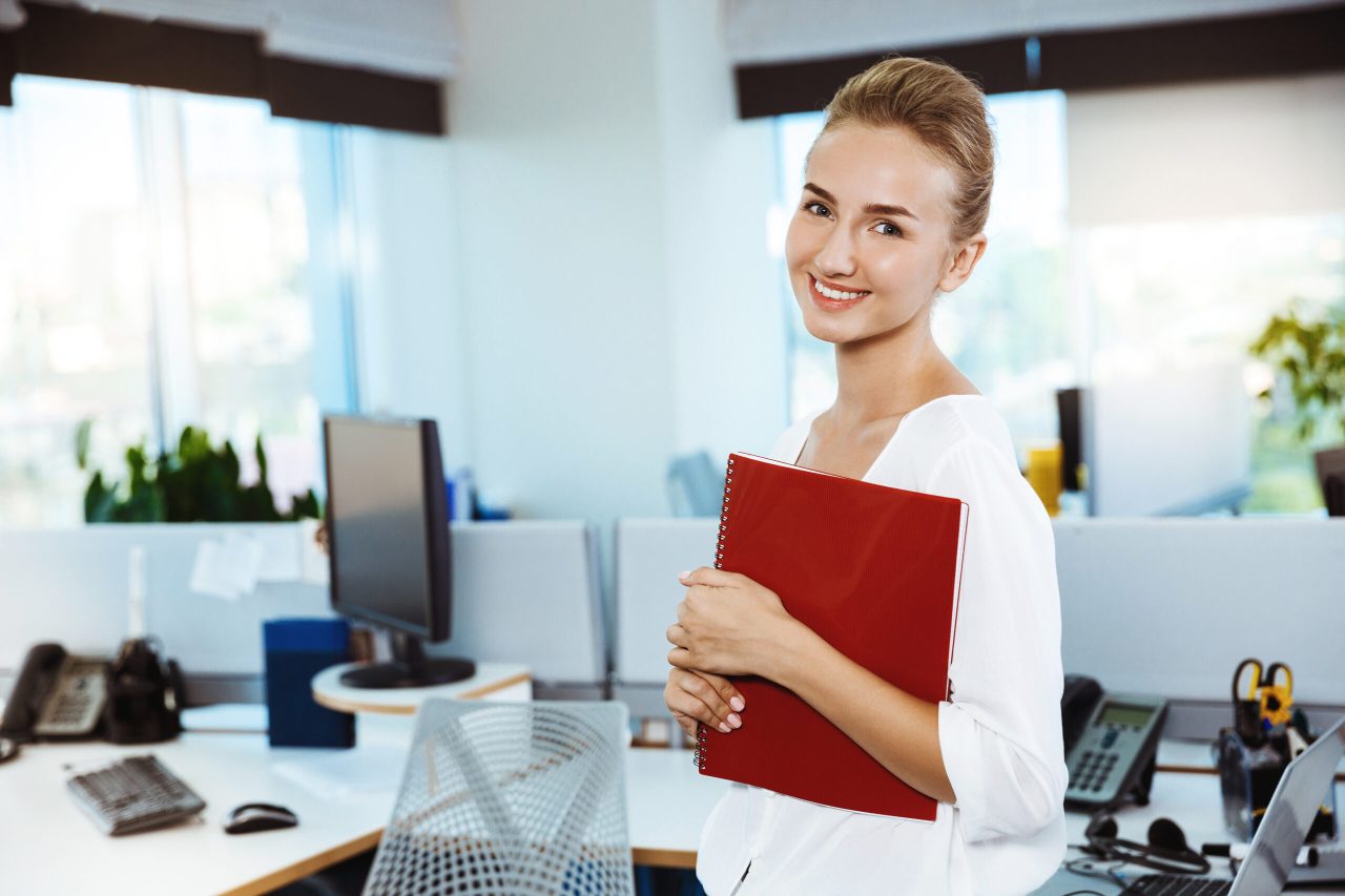 Junge, schöne, erfolgreiche Geschäftsfrau lächelt, posiert, hält eine Mappe, Bürohintergrund